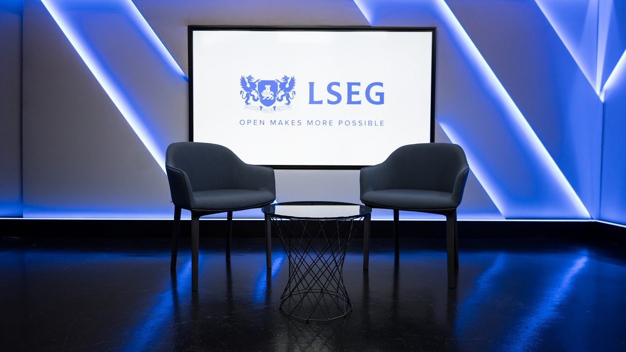 広角撮影したスタジオ 4 で、小型のガラス製コーヒー・テーブルのまわりに椅子 2 脚を配置。ホストの背後のスクリーンに、「オープンであることでより多くのことを可能に」というキャプション付きの LSEG のロゴ。